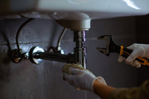Someone in white gloves works on plumbing under sink using wrench