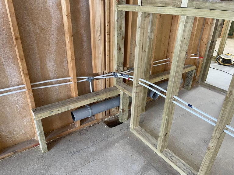 Interior framing of a home addition under construction showing wooden wall studs, metal pipes, and concrete floor