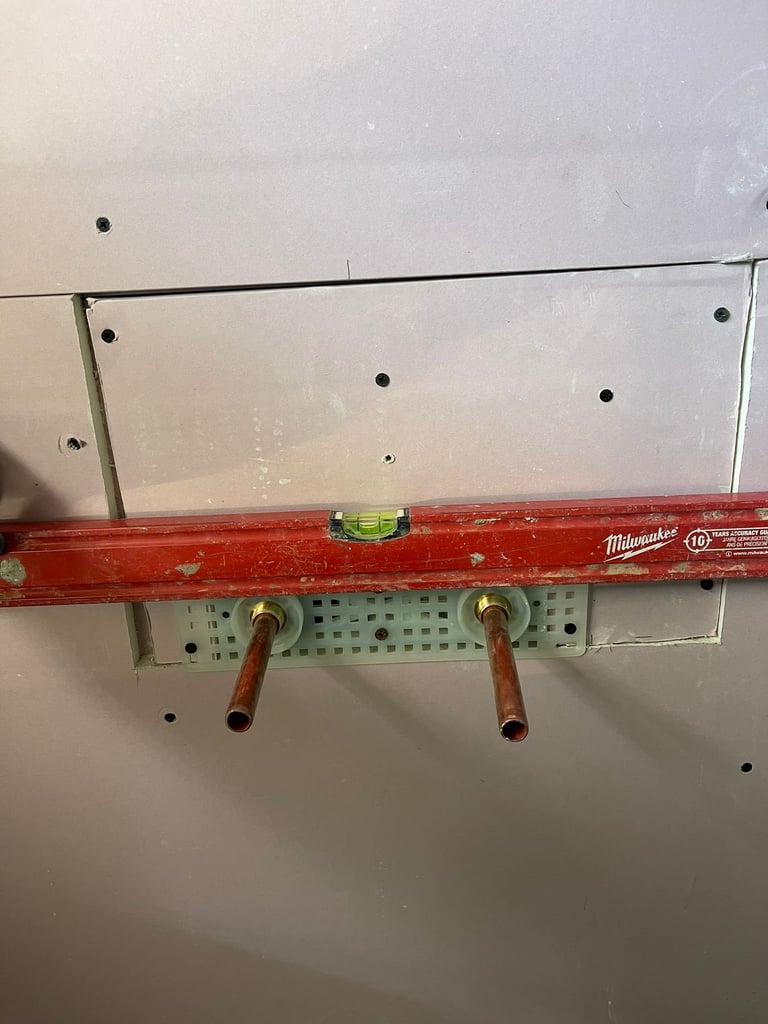 Red Milwaukee spirit level mounted on white drywall with green board underneath and copper pipes below