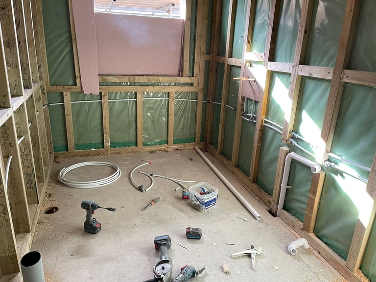Construction site showing a room under renovation with wooden framing, green drywall, scattered tools, and coiled wire on the floor