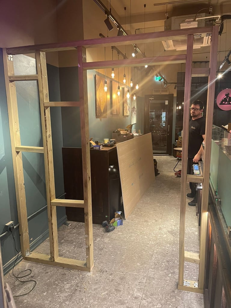 Interior view of a studio or shop space under construction with wooden frame walls, hanging lights, and exposed concrete flooring