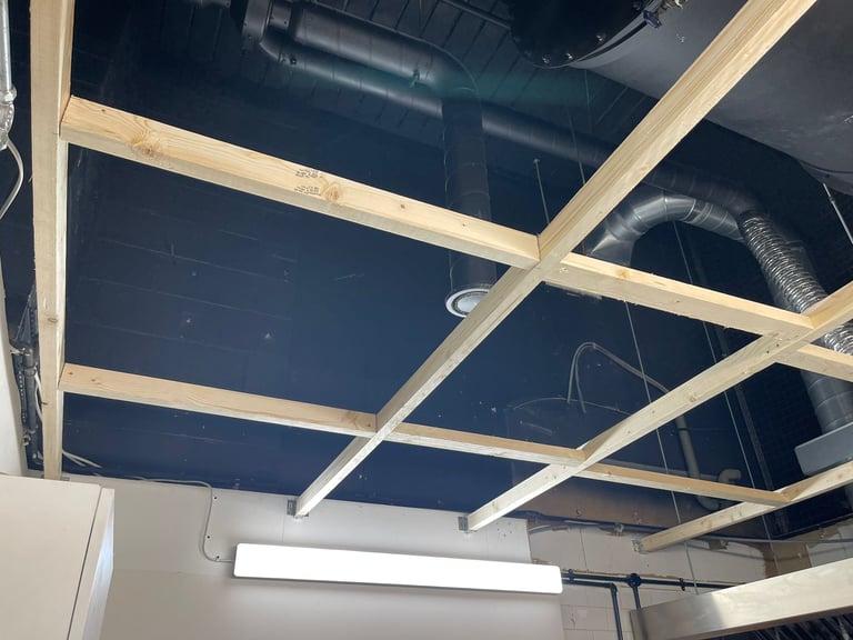 Wooden grid framework mounted on dark ceiling with exposed pipes and ductwork in industrial interior space