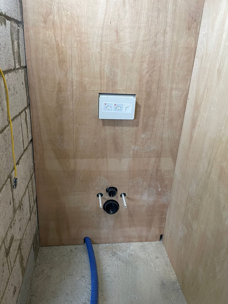 Construction bathroom shower stall with plywood walls, mounted control panel, and black valve fixtures on concrete floor