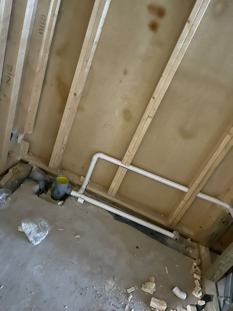 Unfinished basement or utility space with exposed wooden framing, drywall sheets, white PVC pipes, and construction debris on concrete floor