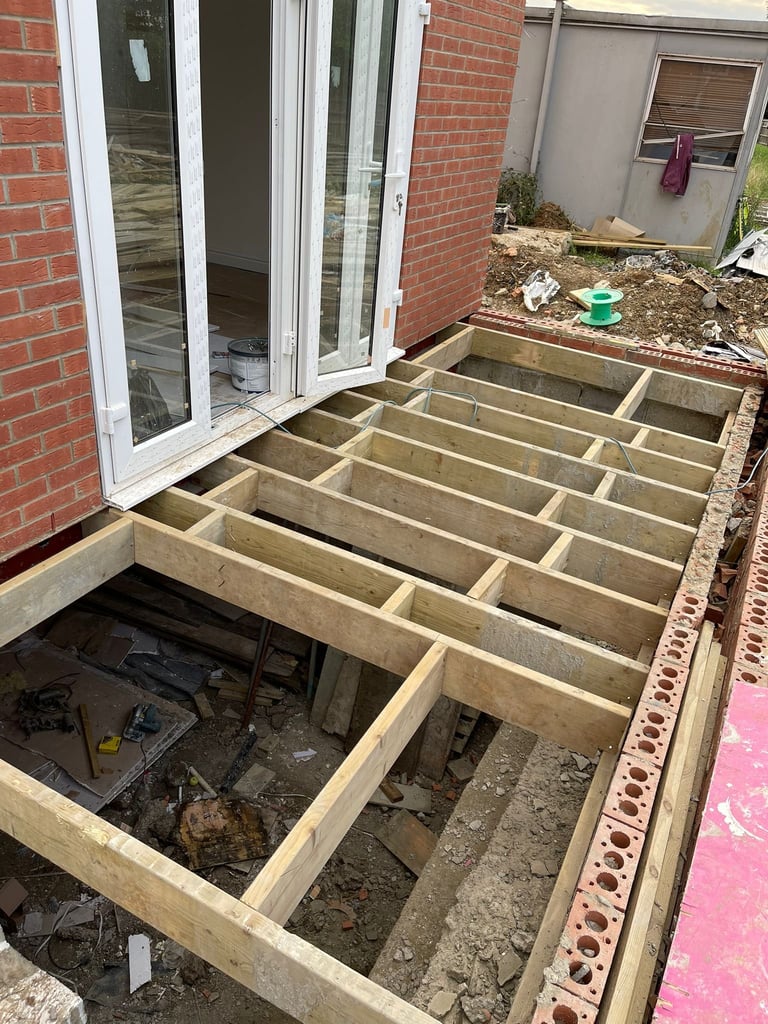 Wooden joists and framework for a deck or patio under construction next to a brick house with glass patio doors