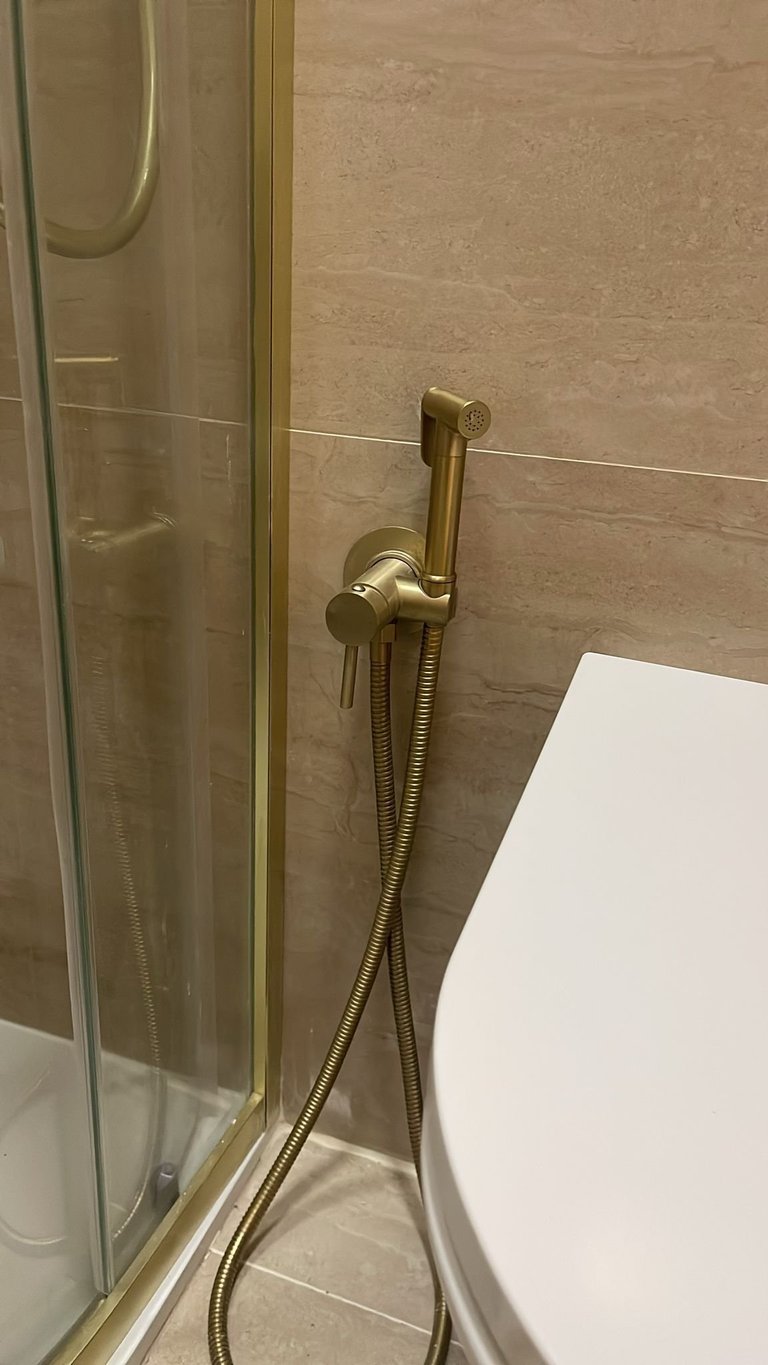 Gold bidet sprayer attached to shower enclosure wall in modern bathroom with beige tile
