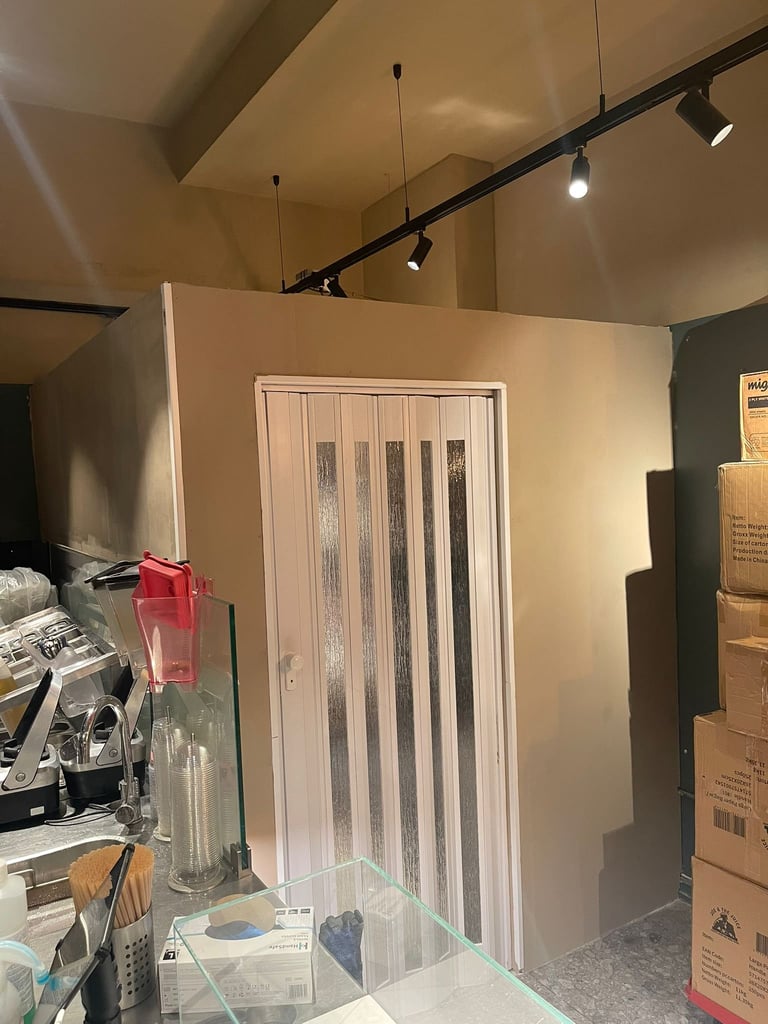 Interior showroom displaying a white accordion-style folding door with vertical panels under track lighting, surrounded by product displays and cardboard boxes