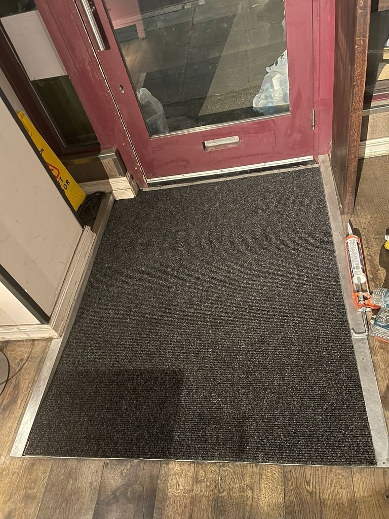 Entry doorway with burgundy door frame, dark gray ribbed entrance mat on wooden floor, and yellow doorstop