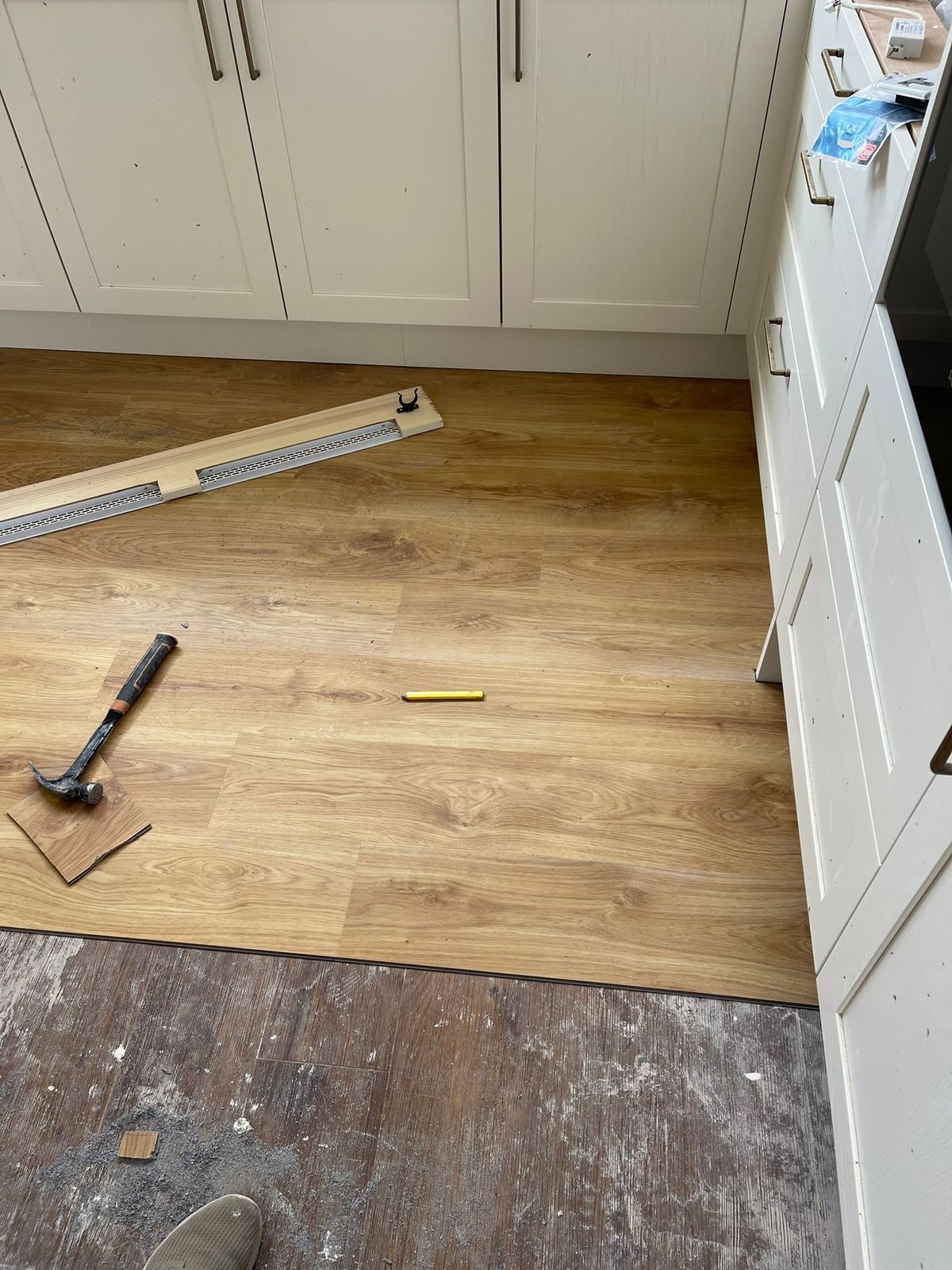 Kitchen floor installation in progress with wood laminate flooring, measuring tape, level, and hammer on light wood surface beside white cabinets