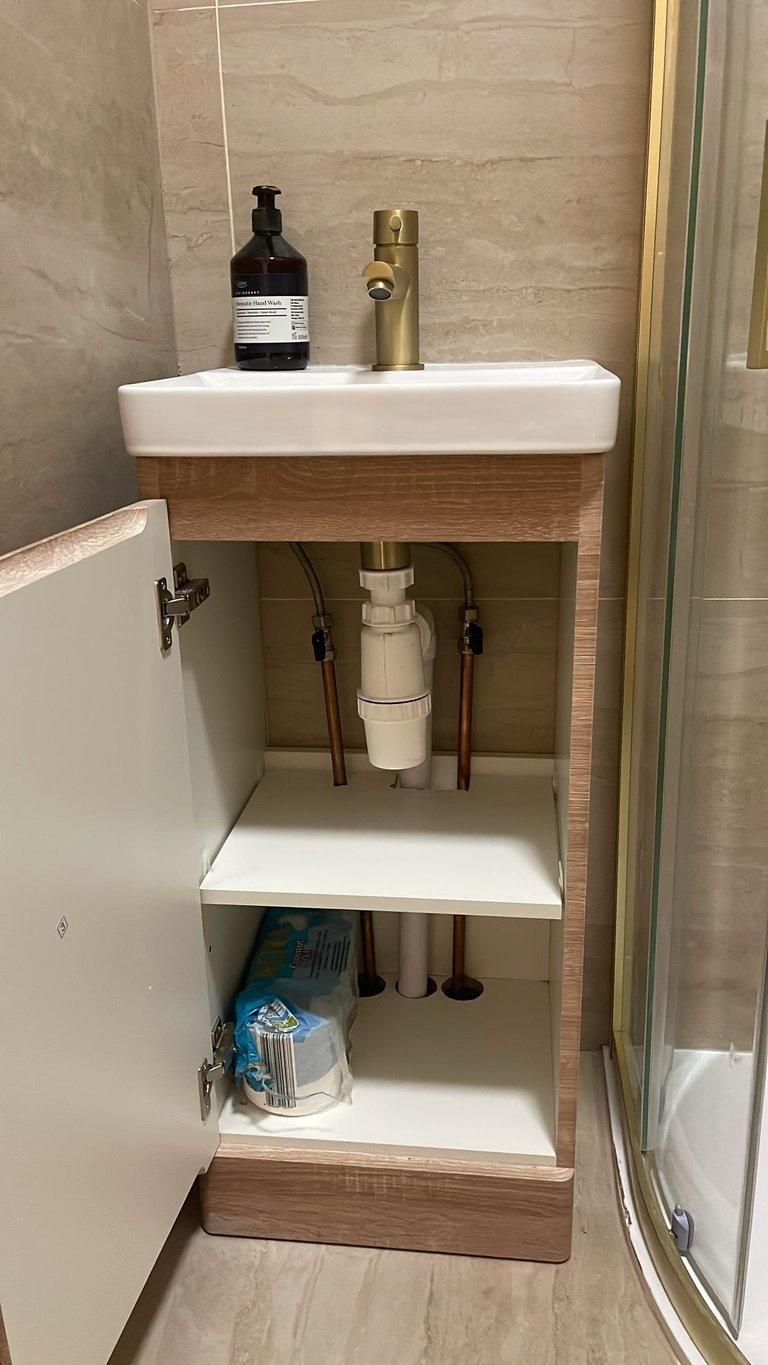 Modern bathroom pedestal sink with open shelving cabinet storing cleaning supplies and toiletries