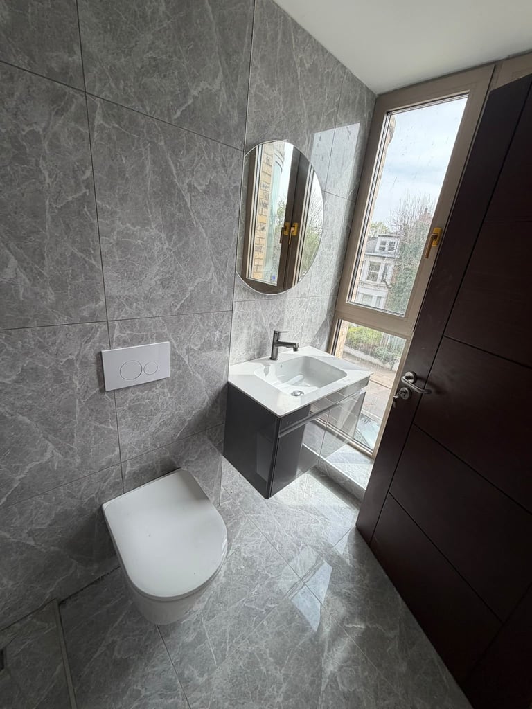 Modern bathroom with gray tile walls, white toilet, wall-mounted sink with mirror, and large windows overlooking a city