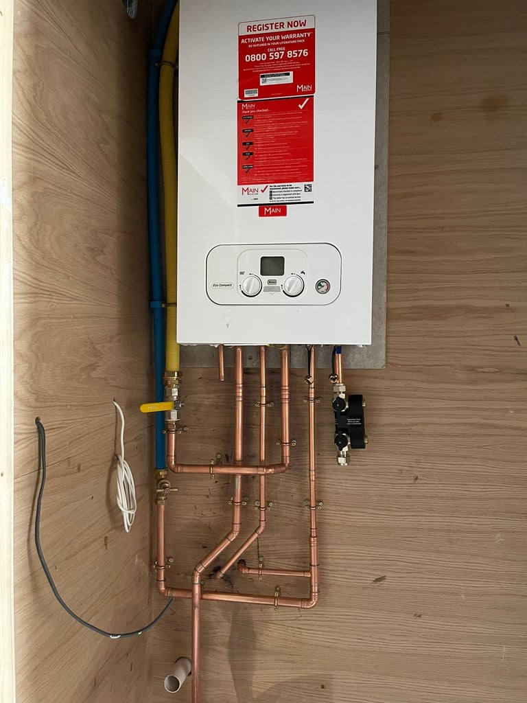 Wall-mounted white boiler unit with digital controls and red registration sticker, connected to copper pipes and electrical wiring on wooden wall
