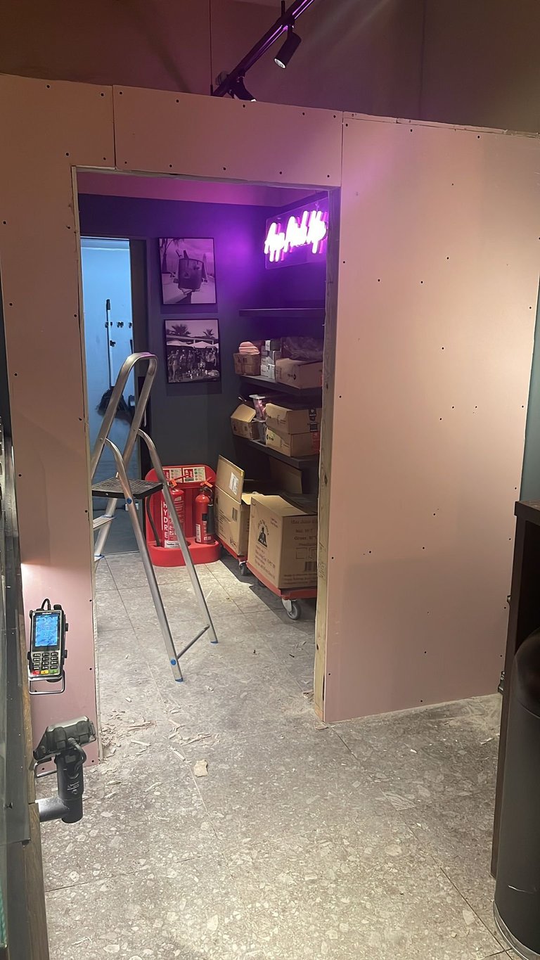 Interior view of a room under construction with pink drywall, purple neon sign, white shelving with boxes, and construction equipment including a ladder and red tool bag