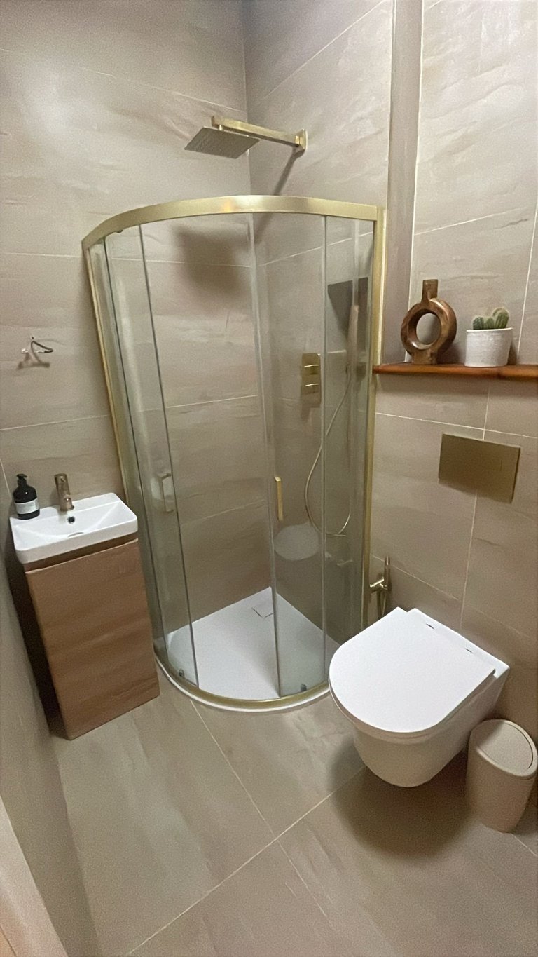 Modern bathroom with curved glass shower enclosure, white toilet, small vanity sink, and beige tile walls