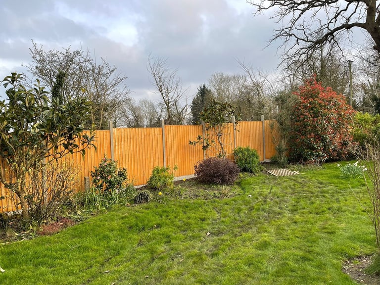 Wooden fence surrounding a residential garden with green grass, bare trees, and shrubs under cloudy sky